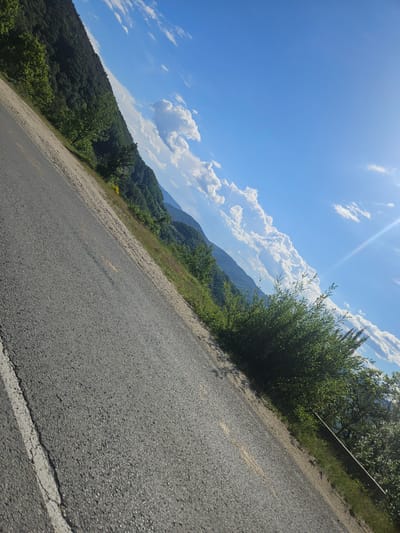 The image shows an asphalt road curving along a hillside in the vicinity of Liaskovo, Bulgaria. To the left of the road is a steep, tree-covered slope, while to the right, vegetation extends up to a metal guardrail. The sky is blue with scattered cumulus clouds. The sun is visible, creating a bright glare. The photograph appears to have been taken during the daytime. There are no people visible in the image.