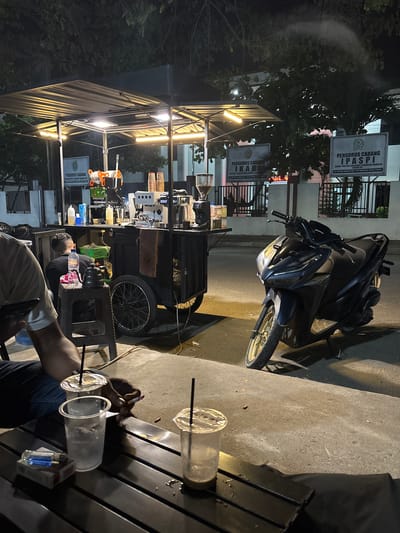 In Gampongjawa, Indonesia, at night, a street-side coffee cart is illuminated by overhead lights. The cart is equipped with a professional espresso machine, coffee grinders, and various containers. A dark-colored motorcycle is parked to the right of the cart. In the foreground, a person's arm is visible, holding a lit cigarette and reaching for a plastic cup containing a beverage with ice and a straw. Another plastic cup with a straw and ice is nearby, along with a pack of cigarettes and a lighter on a dark wooden table. Behind the cart, a person is seated, facing away from the camera, near a collection of items including a kettle and bottles. Two signs are mounted on a fence in the background, one featuring text and a logo for "PENGURUS CABANG IKATAN HAKIM INDONESIA CABANG III" and another for "PENGURUS CABANG IPASPI IKATAN PANITERA SELURUH INDONESIA CABANG ID". Trees and foliage are visible above and around the cart, suggesting an outdoor setting with some greenery.