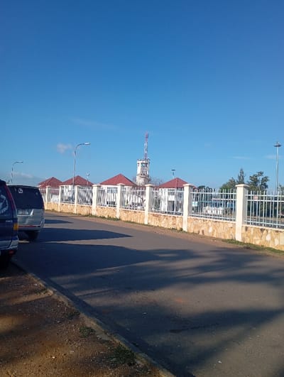 The image presents a daytime street scene, likely in Namanga, Kenya, under a clear blue sky. The perspective is from the side of a paved road, looking forward towards a series of buildings and a prominent tower.
In the foreground, a dark asphalt road dominates the lower half of the frame, with long shadows cast across it from the right, suggesting either early morning or late afternoon sunlight. To the left, a dirt and gravel shoulder with some sparse green vegetation borders the road. Two vehicles are partially visible on the far left. The rearmost car is dark blue, and ahead of it, a silver SUV is seen from behind, with some text on its license plate (e.g., "T 339 D...") being mostly unreadable.
To the right of the road, a decorative fence line runs parallel to the street. It consists of a low stone wall with light-colored, rectangular posts supporting white metal railings. Behind this fence, several single-story buildings with striking red-tiled roofs are visible, interspersed with a few trees. Multiple streetlights, with their lamps on extended arms, are positioned along the street and behind the fence.
Dominating the mid-ground and background is a tall, slender white tower or mast, equipped with numerous antennas and communication equipment, rising above the red-roofed structures. The sky above is a brilliant, cloudless blue, extending across the entire upper portion of the image. The weather appears clear and sunny. No people are visible in the scene. The overall atmosphere is tranquil and brightly lit, characteristic of a pleasant day in the region.