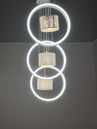 A modern chandelier hangs from a dark ceiling against a light gray wall. The chandelier features three circular rings, each with a decorative lampshade in the center. The lampshades are white with a cut-out pattern of trees and branches, illuminated from within. The rings are glowing white, suggesting they are LED lights. The overall impression is one of contemporary elegance. The image is cropped to show the chandelier from a low angle, with no visible people or specific landmarks of Hanniv, Ukraine.