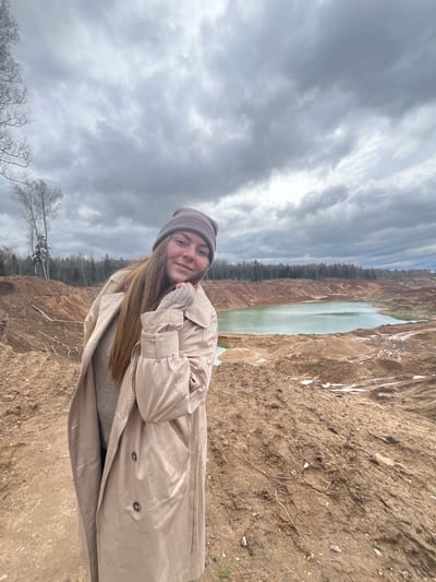 A young woman stands in a landscape of bare earth and sparse vegetation, possibly in or near Ivlevo, Russia. She wears a tan trench coat, a grey sweater, gloves and a beanie, suggesting a cool climate. Her head is tilted slightly, and she looks towards the camera with a soft smile.
Behind her, the ground descends to a narrow lake or pond with greenish water. Bare trees line the opposite bank. Above, a cloudy sky dominates the background. The camera angle is tilted, giving a disorienting perspective of the scene.