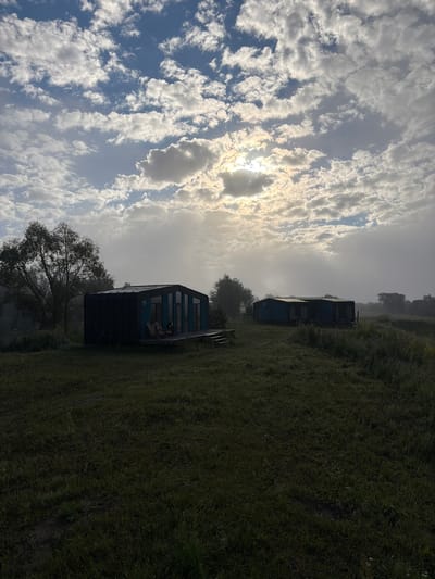 In Avchurino, Russia, a scene unfolds with a modern, dark-colored cabin situated in a grassy field under a cloudy sky. The sun is partially obscured by the clouds, casting a soft glow and creating rays of light. To the left of the cabin, a large tree with dark foliage stands prominently. A wooden deck with two chairs is attached to the front of the cabin, suggesting an outdoor seating area. In the distance, another similar cabin is visible, also nestled within the landscape. The overall environment appears serene and natural, with tall grass and scattered trees contributing to a rural ambiance.