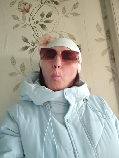 A woman is featured in a close-up, chest-up shot, appearing to take a selfie indoors. She is wearing a light blue puffer jacket with a visible hood and drawstrings, and a light-colored button closure. A white or off-white headband is worn on her head. Her face is mostly obscured by large, rectangular sunglasses with pink-tinted lenses and clear frames. She is making a playful "kissing face" or pout with her lips.
The background shows patterned wallpaper with a floral design, featuring light brown/beige flowers and green/grey leaves on a cream-colored base. A light-colored curtain or drape is visible on the right side of the frame. The setting appears to be an indoor room, likely in a residence. The puffer jacket suggests a preference for warmth. No text is visible in the image. This media file is associated with the location Сусады-Эбалак, Russia.