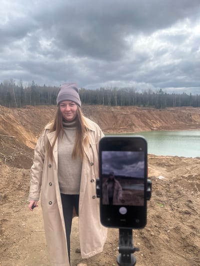 A young woman stands outdoors on a dry, dirt-covered ground, likely in a quarry or excavated landscape, with a large body of water visible in the background. She is wearing a grey beanie, a beige sweater, and a long beige trench coat. She is facing the camera, smiling slightly. A smartphone is mounted on a tripod, positioned in front of her, potentially for taking a picture or recording a video of her in the location. The environment includes steep, earthen cliffs and a dense forest in the background, under an overcast sky. The city of Ivlevo is not discernable from the image.