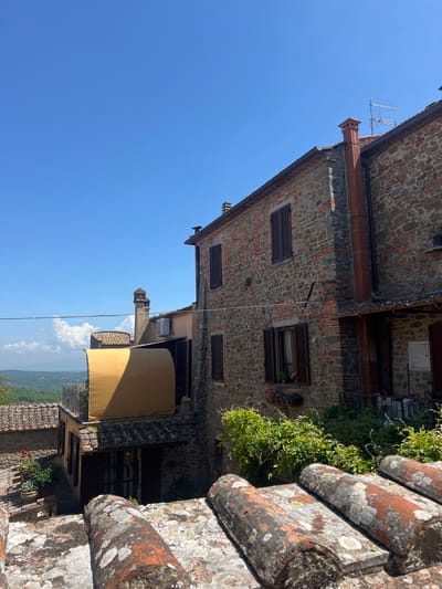 Captured from a slightly elevated perspective, the image showcases architecture within the town of Sinalunga, Italy, set against a clear blue sky. A multi-storied building is constructed of stone with dark-shuttered windows. Patches of greenery are visible between some windows. A light-colored awning is positioned on a lower level. The image's foreground features aged, lichen-covered brickwork, likely part of the observation point. A landscape with mountains is visible.