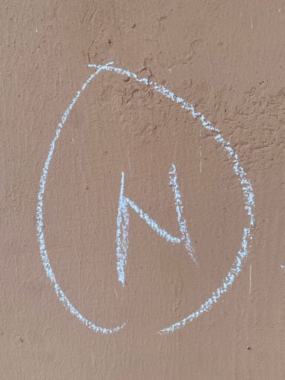 An image shows a chalk drawing of the letter "N" enclosed in a rough oval shape on a textured, light brown surface. The surface appears to be a wall, possibly concrete or stucco, with a slightly uneven texture and some small imperfections. The chalk lines are white and somewhat smudged, indicating they were drawn by hand. The lighting is even, suggesting it was taken in daylight. The location is identified as Dumnga, Nigeria.