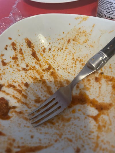 The image displays a close-up, elevated view of a white ceramic dinner plate, post-consumption, positioned on a red surface. The plate's interior is extensively covered with reddish-orange food residue, consistent with a sauce, exhibiting splatters and smears. A four-tined, silver-toned metal fork, featuring a dark handle, rests diagonally across the plate, with its tines oriented towards the lower left. The fork also bears similar food residue. In the upper left background, a crumpled piece of clear plastic wrap is visible. In the upper right background, a partially visible cylindrical Kikkoman soy sauce bottle stands upright. No individuals are present. The scene is an indoor setting in Fegersheim, France.