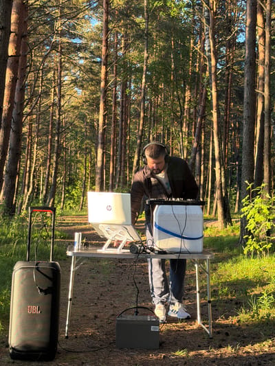 A man wearing headphones is standing behind a table in a forest. He is operating a DJ setup consisting of a laptop, a DJ controller, and a white cooler. A JBL speaker and a battery are also present. The man is wearing a dark jacket, light-colored pants, and white sneakers. The forest is dense with tall trees and sunlight filters through the branches. The ground is covered with pine needles and some grass. The scene suggests an outdoor DJ event or setup in Meremõisa, Estonia.