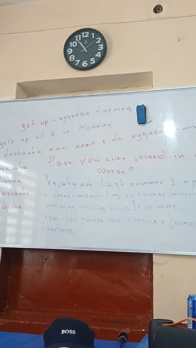 The image shows a whiteboard with handwritten text in English and what appears to be a Central Asian language. A clock on the wall behind the whiteboard reads approximately 10:10. The whiteboard has a blue eraser resting on its top edge. The text on the whiteboard includes phrases like "Get up - uyqudan turmog", "gets up at 6 on Monday", and "Have you ever joined in course?". Further down, there is text about a "three-month English course which improving writing skills. To be honest, I learn from that course how to write a formal structures."
Below the whiteboard, a dark blue baseball cap with "BOSS" embroidered in white is visible. The background appears to be a classroom or meeting room, with a dark brown wooden surface below the whiteboard and a pale yellow wall above. The location is identified as Kattakurgan City, Uzbekistan.