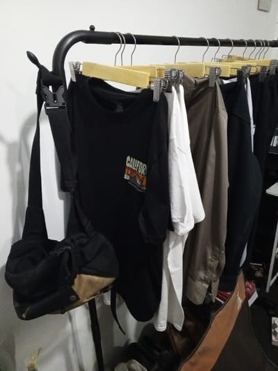 The image displays an indoor scene, located in Al Haouafate, Morocco. A black, tubular metal clothing rack extends horizontally across the frame, positioned against a plain white wall. Several garments are suspended from the rack using a combination of light-colored wooden hangers with metal clips and plain wire hangers.

From left to right, visible garments include a black short-sleeved t-shirt with a "CALIFORNIA" graphic featuring a bridge and skyline in shades of orange and yellow, a white short-sleeved t-shirt, and a long-sleeved, olive-brown or khaki shirt. Other dark-colored clothing items, possibly black t-shirts and trousers, are partially visible further to the right on the rack.

A black and tan fabric messenger or sling bag hangs from the curved left end of the clothing rack. Its black strap is secured by a black plastic buckle, and the bag features a zippered main compartment and a tan lower section with a visible snap closure. The floor beneath the rack is dark, and the lower right portion of the image reveals parts of a brown, possibly leather, object and some indistinct items, including a white structural element, likely part of a shelf or furniture. No individuals are present or engaged in any actions within the scene.