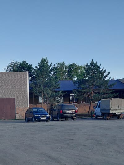 The image depicts an outdoor scene with several vehicles and buildings. In the foreground, a dark blue hatchback car is parked on a gravel surface. To its right, a dark SUV is also parked. Further to the right, a white van with a grey canvas cover over its cargo area is parked. Behind the vehicles, there is a building with a dark blue roof and stone-like lower walls. A tall pine tree stands in front of the building. To the left of the building, another building with a brick-like facade and a brown garage door is visible. The sky is clear and blue. The scene appears to be in a parking lot or an open area adjacent to buildings. The location is identified as Молодёжный, Kazakhstan.