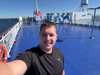 A man is taking a selfie on the deck of a ferryboat under a clear blue sky. The man, wearing a black t-shirt with a white Puma logo, is positioned in the foreground, smiling at the camera. Behind him, the ferry's blue deck stretches out, reflecting the sunlight. In the mid-ground, several passengers are relaxing on deck chairs. Further back, large white and blue shipping containers are visible, adorned with a distinctive blue wave-like pattern. The ferry's superstructure and a red and white funnel rise in the background. To the left of the frame, a bright orange life preserver is mounted on a white railing, and beyond that, the blue water of the sea is visible, with a distant shoreline and a small sailboat. The scene appears to be taking place on the waters near Drottningskär, Sweden, known for its naval history and fortifications.