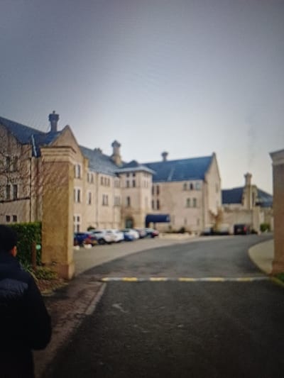 The scene depicts the exterior approach to the Killyhevlin Lakeside Hotel & Lodges, located in Enniskillen, United Kingdom, under an overcast sky. In the foreground, the back of an individual wearing a dark, possibly black, padded jacket is visible in the lower-left quadrant. A paved asphalt driveway, marked with a yellow and black striped speed bump, curves from the lower-center towards the right, leading to the hotel entrance.
The primary subject is a large, multi-story building constructed of light-colored stone or stucco, featuring numerous windows and a dark-colored roof with multiple chimneys. A prominent blue awning or canopy is visible over an entrance area near the center of the building's facade. A line of parked passenger vehicles, including white, blue, and darker colored cars, are discernible in a parking area adjacent to the hotel. Faint smoke or steam is observed emanating from a chimney on the right side of the building. To the left of the driveway, a section of green foliage and bare tree branches is present next to a stone pillar, while a similar stone structure is partially visible on the far right. The overall lighting is diffused, consistent with an overcast day.