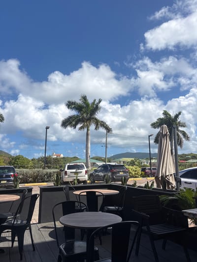 The scene depicts an outdoor dining patio in Spanish Town, British Virgin Islands, under a bright blue sky with scattered cumulus clouds. In the immediate foreground, a wooden deck features multiple unoccupied round tables with dark wood-grain tops and accompanying dark metal chairs. Black planter boxes, containing green foliage, line the edge of the patio. Beyond the patio, a paved parking area is visible, containing a blue SUV on the left, a white SUV in the center, and a dark grey SUV to the right, along with a partially visible white sedan on the far right. Two tall palm trees and two streetlights punctuate the parking lot. A large, folded tan outdoor umbrella stands upright on the right side of the midground. The background consists of verdant, rolling hills with scattered structures, including a distinctive white building with an orange roof on a distant summit. There are no individuals present in the frame.
