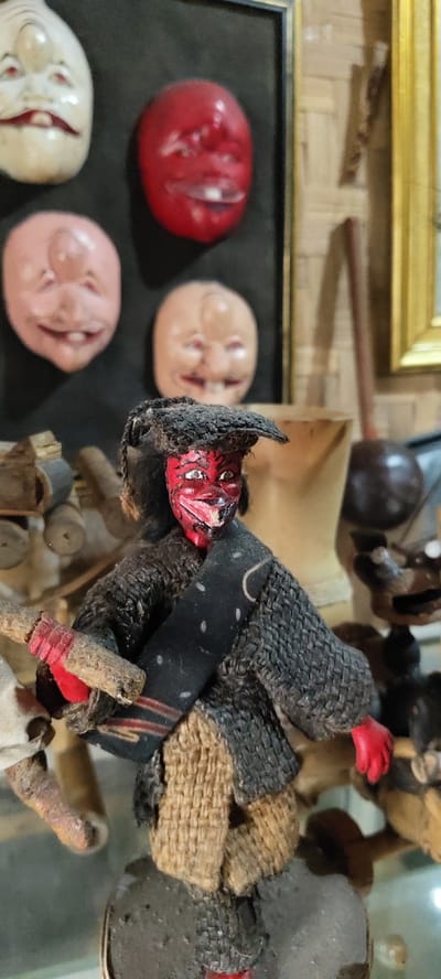 Location: Cikanyere, Indonesia. The scene depicts a close-up, static display of traditional Indonesian artifacts indoors.
In the immediate foreground, a small, dark-skinned doll is prominently featured, standing on a dark, textured base. Its face is painted vibrant red with white eyes and a wide, red-lipped smile exposing visible teeth. The doll wears a dark, woven cap over dark, string-like hair and is dressed in a dark, textured upper garment. A dark sash with red and white patterns is draped across its chest. Its hands are painted red, gripping a rough, light-colored wooden stick horizontally. The doll's lower body is covered in a light brown, coarse woven material.
The midground features various blurred objects. To the left of the doll, blurred light-colored wooden carvings are visible. To the right, a tall, light-colored wooden vessel, possibly a mortar, is present, alongside a dark, rounded wooden ladle or scoop. Further right, additional dark, carved wooden objects are indistinct.
The background primarily consists of a framed display against a dark, possibly black, surface. Within this frame, four distinct mask-like faces are visible: a white, elongated face with a prominent nose at the top left; a bright red, rounded face at the top right; and two light pinkish-peach, rounded faces at the bottom left and right. All mask-like faces feature red eyes and open mouths with visible teeth. To the right of this framed display, a portion of a golden-framed object is visible. The outermost background reveals a light-colored, possibly woven bamboo or rattan wall texture.