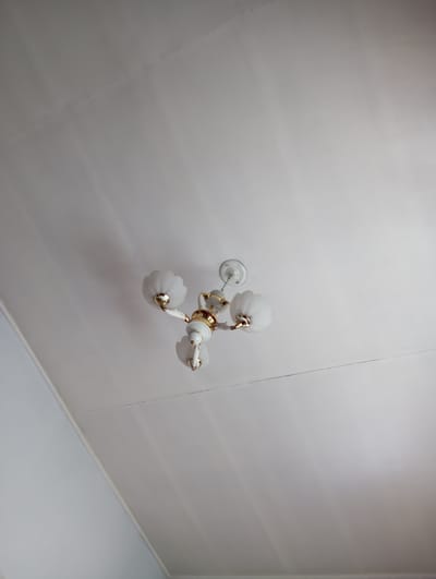 The image shows a white chandelier with gold accents attached to a white ceiling. The chandelier has three arms, each holding a frosted glass lampshade. The ceiling has a visible seam running diagonally across it. The lighting in the room is soft and diffused, casting gentle shadows. There are no people visible in the image. This scene is set in a domestic interior, likely a home or apartment, located in Novonikolaevka, Kyrgyzstan.