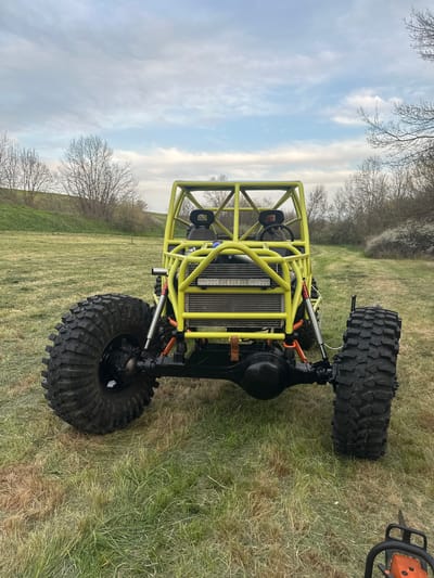 A yellow off-road buggy with large tires is positioned on a grassy surface. The buggy has a custom roll cage and visible radiator at the front. The background features trees, a blue sky, and some clouds. An orange chainsaw is partially visible in the upper right corner. The location is not specified as Choba, Bulgaria within the image.