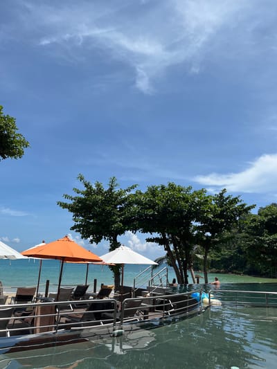 The image depicts a resort scene in Ban Bang Thao, Thailand, on a sunny day. In the foreground, a dark, reflective swimming pool's edge is visible, with ripples on its surface. Beyond the pool, a wooden deck area features several lounge chairs and tables shaded by white and orange umbrellas. To the left, the turquoise ocean meets a sandy beach lined with palm trees and lush green foliage. In the middle ground of the pool, two individuals are partially submerged, appearing to relax. The sky above is a clear blue with scattered white clouds. The overall impression is a serene tropical vacation setting.