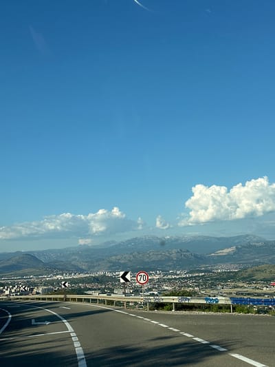 The image depicts a road with a 70 speed limit sign, a city view in the background, a mountain range in the distance, and a blue sky with white clouds. The location is Gornji Kokoti, Montenegro.