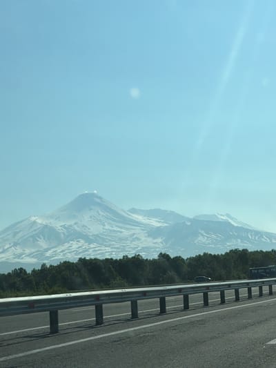 The image captures a scenic view of snow-capped mountains alongside a road in Кеткино, Russia. The scene is dominated by the imposing presence of the mountains, their peaks dusted with white snow, contrasting with the lighter, hazy sky. A dense line of green trees runs along the base of the mountains, separating them from the road.
The road itself is asphalt, with a white line marking the lane. A metal guardrail with prominent posts runs along the edge of the road, separating it from the natural landscape. A dark-colored car is visible on the side of the road, parked near the guardrail, with its hazard lights seemingly on, indicated by a slight orange glow. The car's presence suggests a pause in travel, perhaps for observation or a brief stop.
The lighting suggests it is daytime, with a slightly hazy or overcast sky, casting a soft, diffused light over the scene. The overall impression is one of natural beauty and a road trip through a mountainous region. The image is oriented vertically, with the sky occupying the left half and the road and mountains the right. There is no visible text in the image.