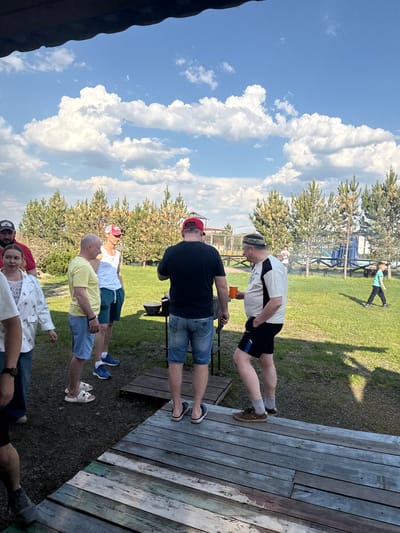 On a sunny day in Кузнецово, Russia, a group of people stand on a wooden platform near a grassy area with trees and a blue sky filled with puffy white clouds. At least six people are visible, dressed in casual summer attire. A man in a white and black shirt and dark shorts stands near the edge of the platform, holding a brown cup. Behind him is a man in a black shirt and denim shorts, and further down the line are people in various colored shirts and shorts. In the background, a person in a turquoise shirt can be seen walking across the grass. A wooden structure with blue accents is also visible in the background.