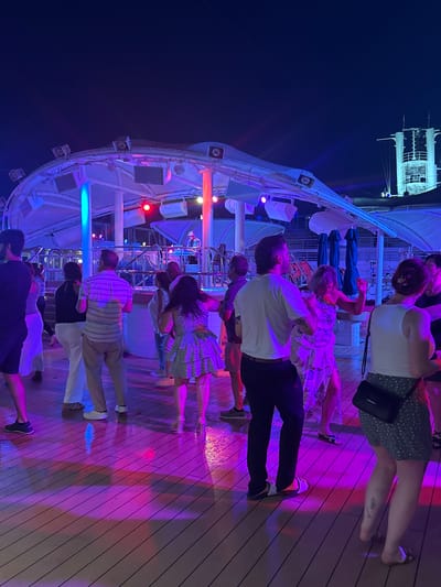 The image depicts a group of people dancing on a wooden deck at night. The scene is illuminated by colorful lights, predominantly purple and blue, casting vibrant reflections on the deck. Some of the individuals are captured mid-dance, with others standing and observing. The background reveals structural elements of what appears to be a ship or a large venue, with lights and railings visible. The overall atmosphere is festive and lively, suggesting an event like a party or a dance gathering. The time of day is clearly night, indicated by the dark sky and artificial lighting.