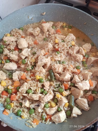 The image shows a close-up, overhead view of a grey speckled pan filled with chicken and rice. The dish contains diced chicken breast, white rice, bright green peas, yellow corn kernels, and diced carrots. Some green beans are also visible. The food appears to be in a savory sauce, and steam suggests it is freshly cooked or being kept warm. The pan is resting on a stovetop, indicated by the dark surface beneath and the edge of a burner visible in the background. The timestamp "27.06.2026 14:39" is overlaid on the bottom right of the image. The setting appears to be a domestic kitchen. The location in the city of Strovolos, Cyprus is not discernible from the image itself.