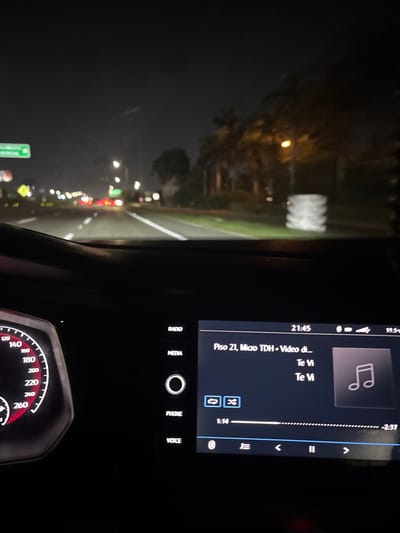 A nighttime view from inside a car. The dashboard is visible with an infotainment screen displaying music information: "Piso 21, Micro TDH - Video di..." and the song title "Te Vi." The screen also shows playback controls and the current time. Below the screen are buttons labeled "RADIO," "MEDIA," "PHONE," and "VOICE." The speedometer is visible, indicating a speed of around 0 km/h. The view through the windshield shows a dark road with streetlights and approaching traffic.