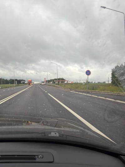 The image shows a perspective from inside a vehicle driving on a road in Kendektamak, Russia. The road is asphalt with double white lines and has streetlights lining the side. The sky is overcast and cloudy, suggesting it is daytime but with poor lighting conditions.

On the left side of the road, there's a no parking sign with a blue circular background and a red X inside a blue circle. Further down the road, there are several vehicles, including what appears to be an orange truck with flashing lights. The scene suggests a typical highway or main road environment. The image appears to be taken through a windshield, with some smudges or water droplets visible on the glass.