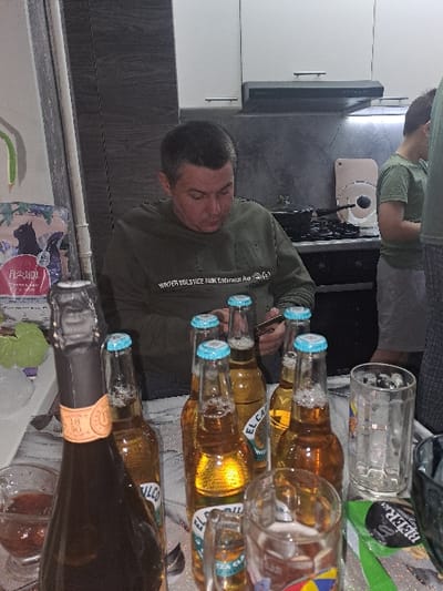 A kitchen counter is cluttered with empty beer bottles and glasses. A man in a green sweater stands by the counter, apparently having consumed some of the beer. Another person is visible in the background. The scene suggests a party or gathering.