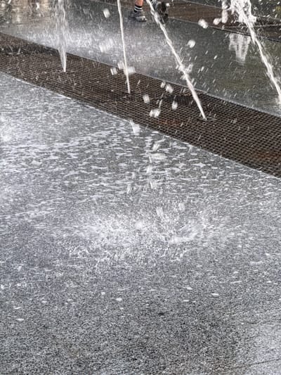 This media file depicts a close-up view of a water feature, likely a fountain, in Piatykhatky, Ukraine. The primary subjects are water and the textured surfaces it interacts with. The scene is dominated by a diagonal band of dark, metallic grating that runs across the left side of the frame, with water spraying from it and onto a speckled, gray stone surface on the right. The stone appears to be granite, characterized by its granular texture and variations in gray tones.
The activity taking place is the spraying of water. Streams and droplets of water are visible in various states of motion – some are sharp, jet-like streams emanating from the grating, while others are dispersed into a fine mist or are captured as splashes and droplets on the surfaces. The water creates a dynamic and somewhat abstract pattern against the solid backgrounds.
Notable details include the contrast between the uniform gray of the stone and the dark, geometric pattern of the grating. The play of light on the water causes some areas to appear bright white, highlighting the movement and texture of the spray. The time of day and weather are not explicitly indicated, but the bright lighting suggests it is daytime with clear conditions. No emotions are discernible as the subjects are inanimate. There is no visible text in the image. The context of Piatykhatky, Ukraine, provides a geographical reference for the location of this water feature.