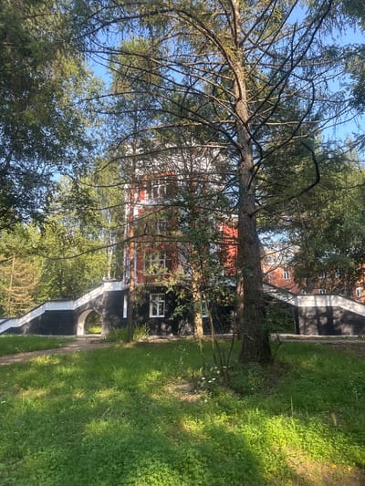 The image captures a building with red and white facade, partially obscured by trees. A black and white arched structure leads to the building. The foreground is a grassy area with sunlight filtering through the trees. The location is the sanatorium "Ust-Kachka" in the Perm Krai, Russia.