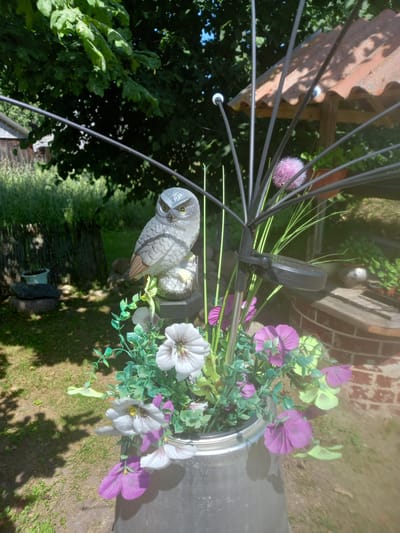 The image shows a close-up of decorative items in an outdoor setting, likely a garden. The main subjects are two owl figurines, one larger and more detailed than the other, positioned in front of artificial flowers. The larger owl is grey with distinct feather patterns and yellow eyes, and it appears to be perched on something. Surrounding the owls are artificial flowers in shades of purple, light yellow, and white, with green leaves and stems. These flowers are arranged in a metallic container, possibly a bucket or planter.
Behind the decorative elements, glimpses of the surroundings can be seen. There is a reddish-brown tiled roof on the left, and a brick structure in the background to the right. Lush green foliage from trees or bushes forms a backdrop. The ground is covered in grass and some patches of dry earth. The lighting suggests it is daytime, with sunlight casting shadows, indicating a sunny day.
The overall impression is of a decorated outdoor space, possibly a garden or patio, with the owl figures and artificial flowers serving as ornamentation. There is no activity or event taking place, and no people are visible. The scene is static and focuses on the decorative elements and their immediate surroundings. No visible text is present in the image. The location context of Adutiškis, Lithuania, suggests a possibly rural or suburban setting.
