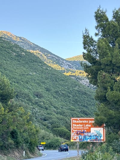 The image shows a mountain landscape in Medinski Krš, Montenegro, with a road winding along the side. A sign indicates Skadar Lake National Park, defaced with graffiti. Two vehicles are visible on the road. Dense vegetation covers the hillside.