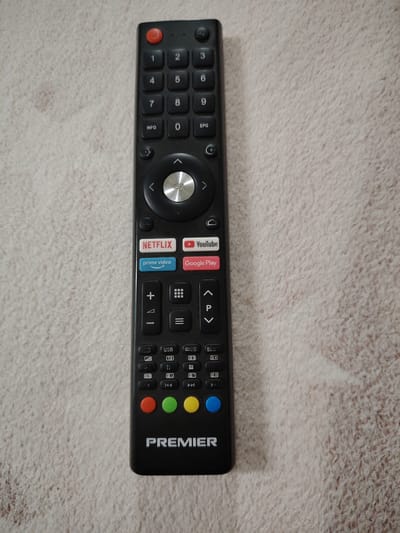 A black remote control with a textured, light-colored background is pictured. The remote has a power button at the top, followed by a numeric keypad with buttons labeled 0 through 9, and additional buttons for INFO and EPG. Below the numeric keypad is a directional pad with an OK button in the center. Further down are dedicated buttons for Netflix, YouTube, Prime Video, and Google Play. A plus and minus button, as well as up and down arrows, are visible, along with other function buttons. The bottom of the remote features four colored circular buttons (red, green, yellow, blue) and the brand name "PREMIER". The background appears to be a woven fabric or carpet. There is no indication of the city of Bokhtar, Tajikistan in the image.