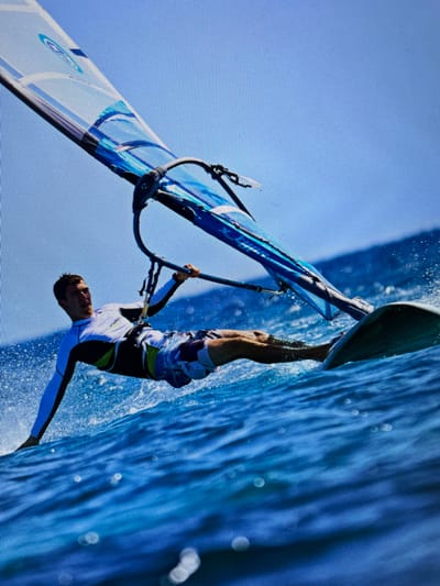 A man windsurfing is upside down on the water. He is wearing a white wetsuit with dark sleeves and swim trunks. The water is blue with white streaks. The sky is light blue. He is holding onto the boom. The sail is white and blue. This is not taking place in Bacong, Philippines.