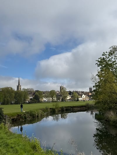 The image, when correctly oriented, captures a tranquil daytime scene of a village or town situated along a body of water, likely a river or canal. The sky is partially covered with soft grey clouds, revealing patches of clear blue sky.
On the left side of the frame, a wide, vibrant green grassy bank stretches along the water. A person, dressed in dark clothing, is visible walking along this bank, accompanied by two light-colored dogs. Behind the bank, a collection of traditional stone buildings forming a village or town are visible, nestled among trees. At least two distinct church spires or towers rise above the rooftops, indicating significant historical architecture. Various trees, some with lush green foliage and others with more sparse branches, punctuate the landscape along the bank and among the buildings.
The right side of the image is dominated by the calm, reflective surface of the water, which perfectly mirrors the cloudy and blue sky, as well as the structures and trees from the opposite bank. The reflections are remarkably clear, creating a sense of depth and symmetry. In the immediate foreground on the right, some wild grasses and white flowers grow along the water's edge. A wooden post or bench is partially visible in the lower-left foreground.
The overall atmosphere is peaceful and serene, suggesting a pleasant day, likely during spring or early summer given the fresh greenery. There is no visible text in the image. The architectural style and natural setting are characteristic of a rural or semi-rural location in the United Kingdom.