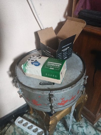 The scene, located in the city of Dogsenang Gorphoog, Bhutan, depicts a static arrangement of objects in an indoor environment. A weathered, silver-grey snare drum, exhibiting red paint chips and surface abrasions, rests on a decorative gold-colored, possibly wooden, three-legged stand. Positioned atop the drumhead is an open, empty black cardboard box with visible text "DEMONIK" and "4000GRIT," alongside a rectangular package of "Yoa 100% NATURAL BAMBOO PREMIUM FACIAL TISSUE," featuring a panda graphic and green accents. To the bottom left, on a light green patterned floor, a white five-socket power strip is visible. The background consists of a light-colored wall with a white cable running vertically, and a dark brown wooden furniture item or shelf is partially visible on the right. No individuals are present.