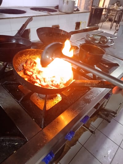 In a commercial kitchen located in Kasri Arifan, Uzbekistan, a dark, round-bottomed pan, containing multiple pieces of seared meat or poultry, sits atop an active gas burner on a stainless steel commercial stovetop. A large, bright orange-yellow flame erupts from the pan, enveloping its contents. A human hand, visible on the right, firmly grips a long-handled metal ladle, which is partially submerged within the flaming pan, indicating an ongoing cooking action such as stirring or flambéing. The stainless steel stovetop features visible blue control knobs on its front panel. In the background, several other large, dark-colored pans, one with a long wooden handle, are positioned on adjacent burners or resting on the counter. A smaller, dark pot, a metal colander, and another ladle are also visible on the stainless steel counter surface. Further back, a white tiled backsplash delineates an elevated counter holding two large, lidded metal containers. Distant, out-of-focus dark tables and chairs suggest an adjacent dining or storage area. The floor beneath the stovetop is composed of white tiles, showing some moisture and general wear.