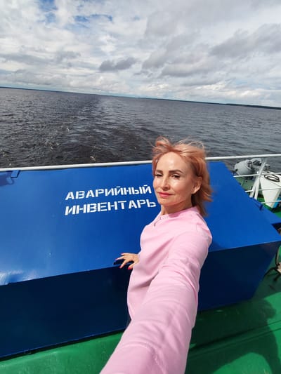 A woman with blonde hair and pink clothing is taking a selfie on the deck of a boat. She is standing in front of a blue container with white Cyrillic lettering that reads "АВАРИЙНЫЙ ИНВЕНТАРЬ" (Emergency Equipment). The boat is on a large body of water, likely a river or lake, with a cloudy sky above. The background shows the water stretching to the horizon, with a faint line of trees or land visible in the distance. The image appears to have been taken during the day, with sunlight illuminating the scene. The location is identified as Kostyaevka, Russia.