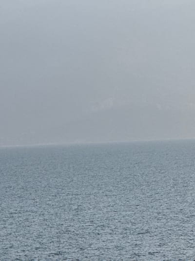 The image displays a seascape from Kato Karioutes, Greece, presenting a horizon line that divides a body of water from an atmospheric sky and distant land. The lower one-third of the frame is occupied by water, characterized by small, textured ripples across its surface, appearing in a muted blue-grey hue. The upper two-thirds of the frame consists of a light grey, hazy sky, through which indistinct, darker outlines of distant landmasses or mountains are faintly discernible. No individuals, vessels, or specific objects are identifiable within the scene. The environment is natural, exhibiting reduced clarity due to atmospheric conditions.