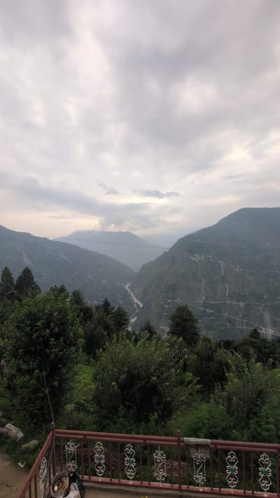 An outdoor, eye-level, vertical shot captures a panoramic view of a valley in Cheelaage, India, under a cloudy sky. The foreground features a dark red railing with white ornate scrollwork, a trash bin, and some plastic bags. Behind the railing, dense green foliage creates a natural border. Beyond the vegetation, the landscape unfolds into a vast valley with lush green slopes dotted with scattered settlements. A winding river snakes through the valley floor, reflecting the muted light of the overcast sky. The mountains recede into the distance, their peaks shrouded in a hazy atmosphere. The overall impression is one of serene natural beauty and the quiet presence of human habitation within a majestic landscape.