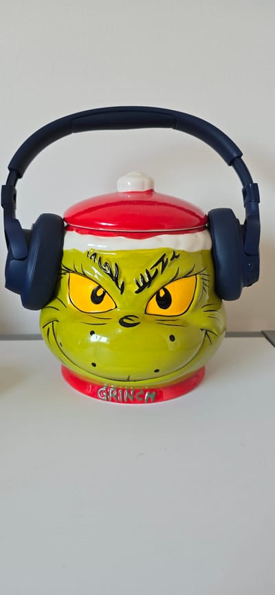 A bright green Grinch cookie jar with large yellow eyes, black eyebrows, and a red and white Santa hat is displayed on a white surface. The cookie jar is wearing a pair of dark blue over-ear headphones. The word "GRINCH" is written in metallic letters on a red collar around the base of the jar. The background is a plain off-white wall. No specific location in Drummondville, Canada is discernible from the image.