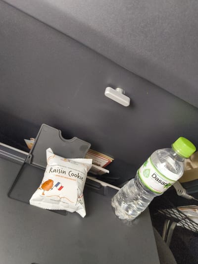 In a dark, enclosed space, possibly the interior of a vehicle or an aircraft, a bottle of Dasani bottled drinking water with a green cap rests on a black surface. Next to it, a packaged "Raisin Cookie" with French imagery, including a flag and the Eiffel Tower, is visible. A grey plastic tray holder is also present, containing some papers. Above these items, a silver-colored latch is mounted on a textured grey surface. The overall scene suggests a travel environment. The image is not sufficient to determine the location in Kon Sỏ, Vietnam.