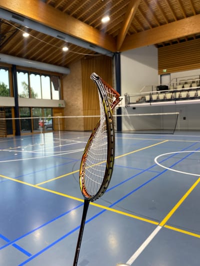 The image shows a warped badminton racket held in the foreground, with a badminton court in the background. The court has blue flooring with yellow and white lines marking the boundaries. Two badminton nets are visible, one in the mid-ground and another further back. Spectator seating is visible in the upper right background. Large windows are present on the left side of the image, revealing trees and a parking lot outside. The ceiling of the indoor sports facility features exposed wooden beams. The scene appears to be indoors at the Centre sportif, Rue des Maladières 11, 2016 Cortaillod, Switzerland. There are no people visible in the image.