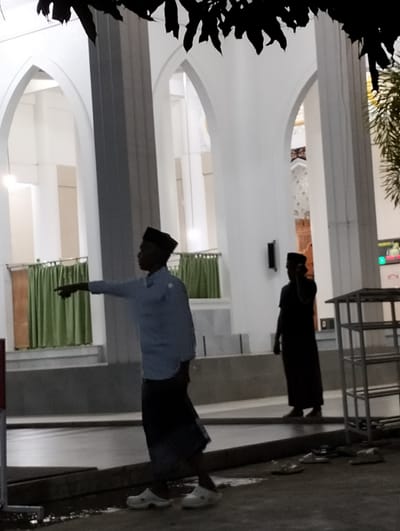 The image depicts an outdoor scene at dusk, likely in Blangmetimu, Indonesia, with elements suggesting a mosque or religious building. In the foreground, a man wearing a light blue shirt and a black cap is pointing to the left. He is also wearing a dark skirt. To his right and slightly behind him, another man in a black outfit and black cap is standing, partially obscured. The background features white architectural elements with arched openings, reminiscent of mosque design. Green curtains hang behind a railing. A metal shelving unit is visible on the right side of the image, and several pairs of sandals are scattered on the ground in the lower right corner. The overall lighting is dim, characteristic of evening.