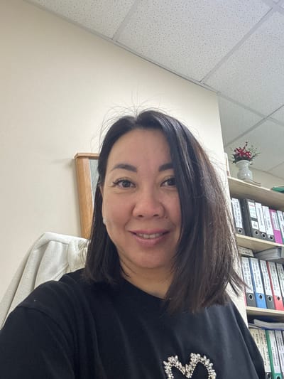 The image shows a medium shot of a woman in an office setting. She has dark brown hair, parted in the middle and falling to her shoulders. She is wearing a black t-shirt with a sparkly embellishment on the chest. She is smiling and looking directly at the camera. In the background, there is a wall, a ceiling with acoustic tiles, a shelf filled with binders, and a small decorative plant in a vase. To the left of the woman, a light-colored jacket or coat is draped over a chair. The overall impression is that of a casual selfie taken in an office. The image does not provide any specific details about the location within the city of Yakty-Kul, Russia.