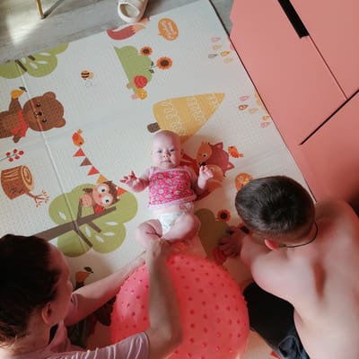 In Trubacheyevka, Russia, on a soft, printed mat designed with cartoon animals and nature motifs, a family is engaged in playtime. A baby, wearing a pink strawberry-patterned top and a diaper, lies on its back, looking up with wide blue eyes. Two adults are positioned around the baby. A woman, with reddish-brown hair and wearing a pink t-shirt, is on the left, her arms reaching towards the baby. A man, with dark hair and bare-chested, is on the right, leaning over the baby. A large, spiky pink ball is centrally located on the mat, with the baby's legs resting on it and the adults' hands interacting with it. The background shows a portion of a pale wooden floor and a tall, peach-colored cabinet. The lighting suggests an indoor setting, possibly a living room or playroom.