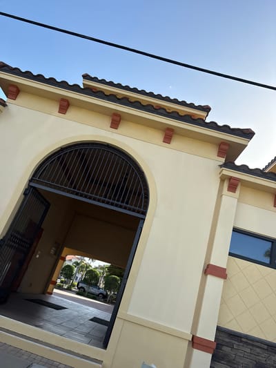 A tilted, exterior, medium-shot perspective captures a section of a building's archway entrance in Punta Gorda, USA. The building is painted a light yellow with terracotta accents along the roofline and structural supports. The entrance features a large, arched opening with a black wrought-iron gate partially open, revealing a glimpse of a walkway and some greenery. A dark power line is visible cutting across the bright blue sky.