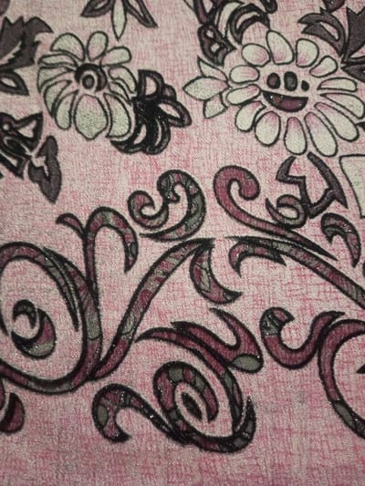 A close-up reveals a pink textile with a floral and swirl pattern. Flowers with white petals and dark centers are intermixed with dark swirling tendrils outlined in black. The background has a textured look, with fine lines or a crosshatch pattern. The swirl elements appear in shades of dark red.